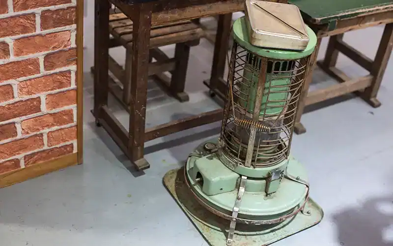 Een vintage petroleumkachel die kachelbrandstof gebruikt, geplaatst op een grijze vloer naast een houten tafel en stoel. De kachel is groen met metalen beschermroosters en staat op een metalen plaat.