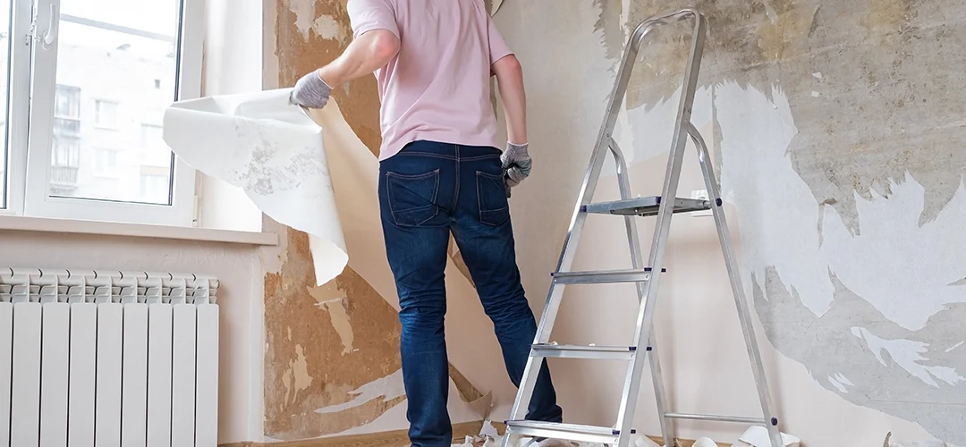 Man verwijdert oud behang van de muur met de hand, naast een trap in een kamer tijdens renovatie.