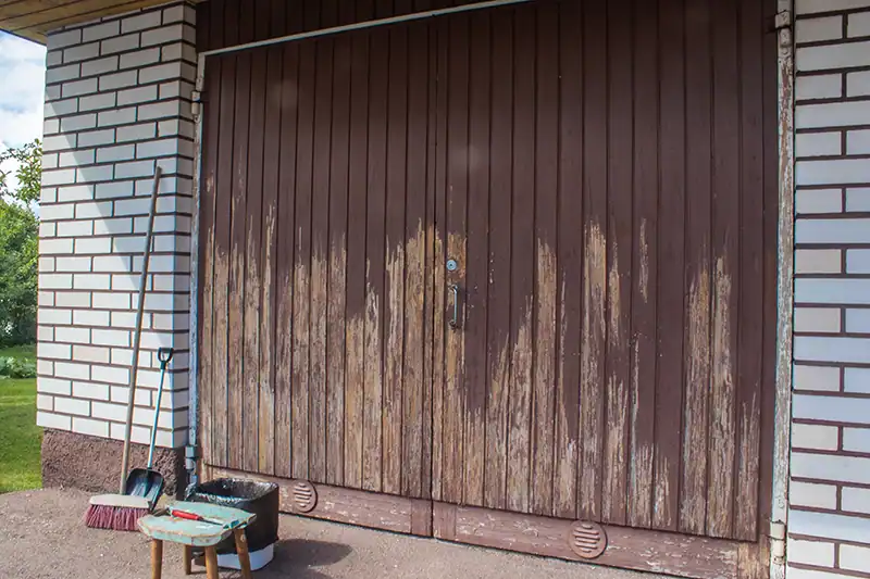 Wooden garage doors with partially peeling paint and prepared cleaning supplies beside them.