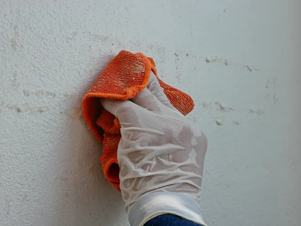 A hand wearing a plastic glove uses an orange cloth to remove adhesive residue from a white wall surface.