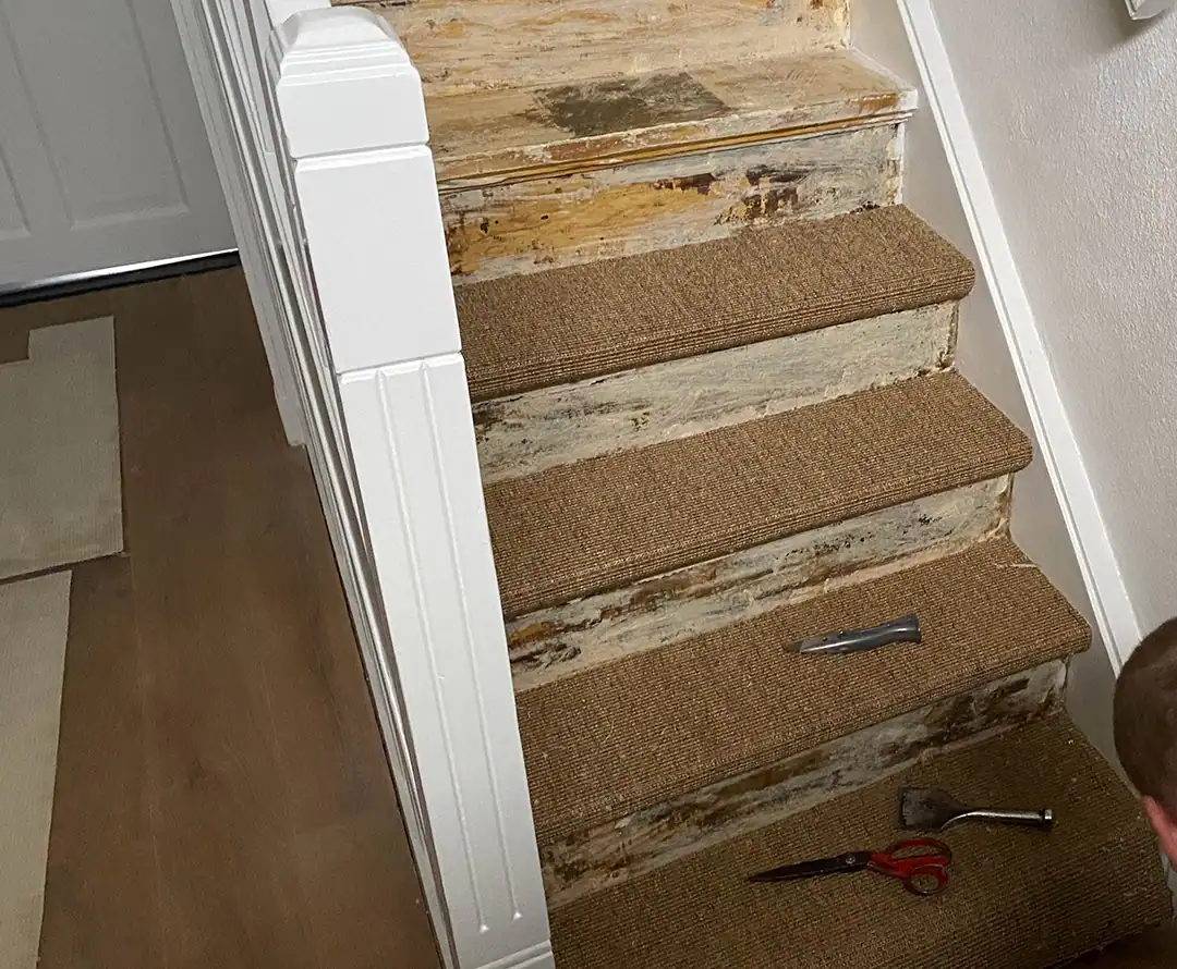 An image of a wooden staircase with the carpet being removed, showing adhesive residue and tools such as a utility knife and scraper on the steps.